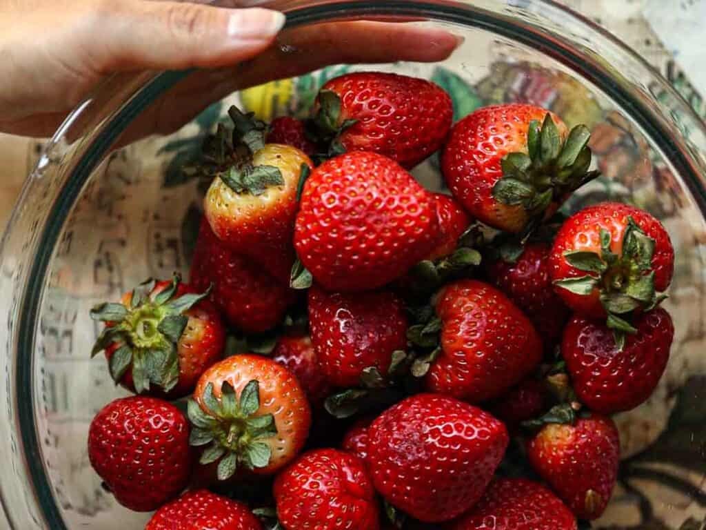 A hand holding a glass bowl filled with bright red strawberries, some with leaves still attached. The background features a colorful, patterned surface.